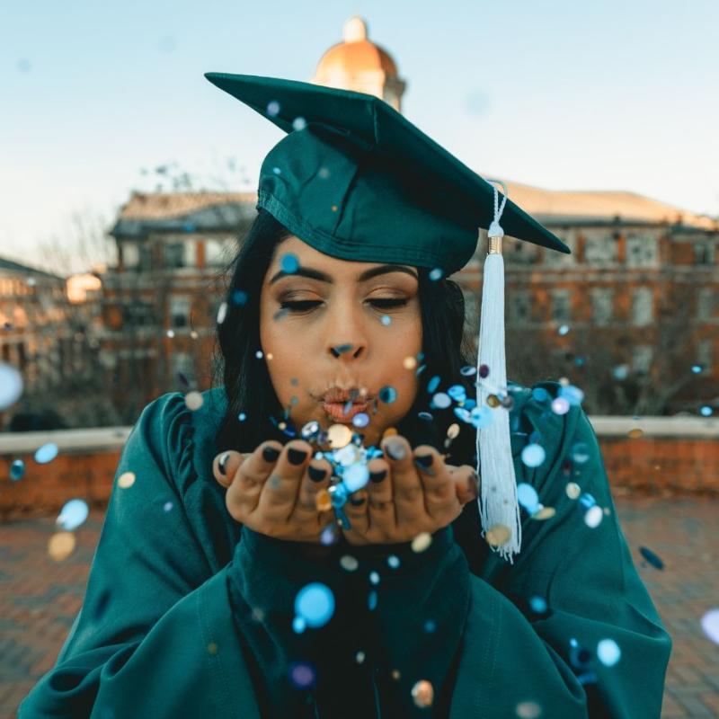 Girl in green cap and gown blowing confetti out of her hands